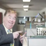 Man in a suit smiling and pointing at a glass award inside an office with cubicles.