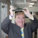 Person in a suit holding up a clear award trophy, smiling in an office setting.