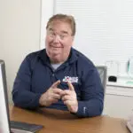 A person sitting at a desk, gesturing with their hands, wearing a navy blue shirt. A computer monitor is partially visible in the foreground.
