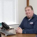 A man in a blue shirt sits at a wooden desk with a computer and phone, smiling, in an office with a window.