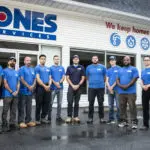 A group of ten people in blue shirts stands in front of a building with a "Jones Services" sign and various service icons.