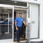 A man in a blue shirt and cap stands at the open glass door of a storefront.