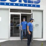 Two people in blue uniforms stand outside a building with a glass door and a sign reading "SERVICES.