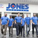Six people in matching blue shirts stand in front of a building with a "Jones Services" sign.