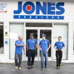 Four individuals in blue shirts walk out of a building with "JONES SERVICES" signage above the door.