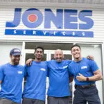 Four people in blue shirts stand smiling in front of a building with a "JONES SERVICES" sign.