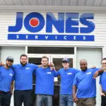 Six people in blue uniforms stand smiling in front of a building with a sign reading "Jones Services.