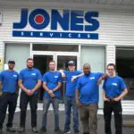Six people wearing blue shirts stand in front of a building with a "JONES SERVICES" sign.