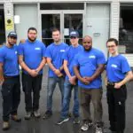 A group of six people wearing blue shirts stands in front of a building with glass doors.