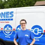 Person in a blue uniform stands smiling in front of a service van displaying plumbing, heating, and electric symbols.