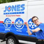 Person in a blue uniform smiling and pointing at a van labeled "Jones Services" with plumbing, heating, and cooling symbols.