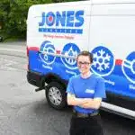 Person in a blue uniform standing with arms crossed in front of a white van with colorful graphics and company logo.