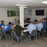 People sitting around tables in a meeting room, some wearing blue shirts. A whiteboard and TV are mounted on the walls.