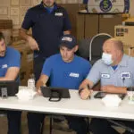 Four men in blue shirts, one wearing a mask, are seated at a table with tablets in a room filled with boxes. Another man stands behind them.