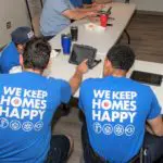 Two people in blue shirts with "We Keep Homes Happy" sit at a table, looking at electronic devices.