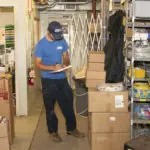 A person in a blue shirt writes on a clipboard in a storage room with shelves and boxes.