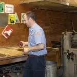 Person in a blue shirt standing at a workbench in a workshop, with tools and safety signs visible on the wall.