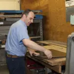 A person in a blue shirt works with materials on a wooden table in a workshop. Shelves and tools are visible in the background.