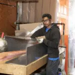 A person wearing glasses and gloves stands next to a large metal structure in a workshop, holding a tool.
