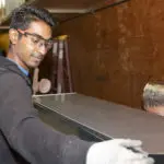 A person wearing safety glasses and gloves works with a metal sheet, focusing intently on the task. A ladder and wooden surface are visible in the background.