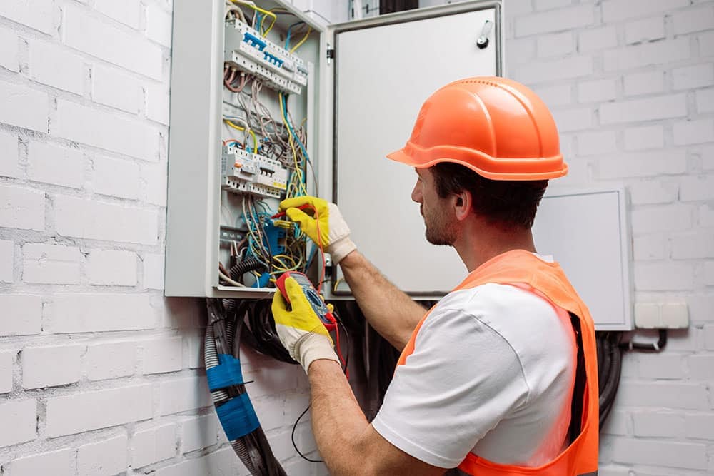 An electrician wearing an orange hard hat and vest inspects and repairs wiring inside an electrical panel on a white brick wall, carefully diagnosing electrical noise to ensure safe operation.