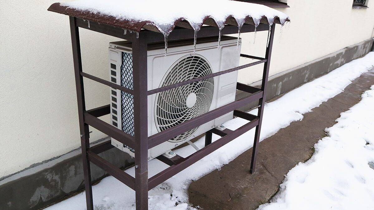 Outdoor air conditioning unit sheltered by a metal frame, with icicles hanging from the roof and snow covering the ground.