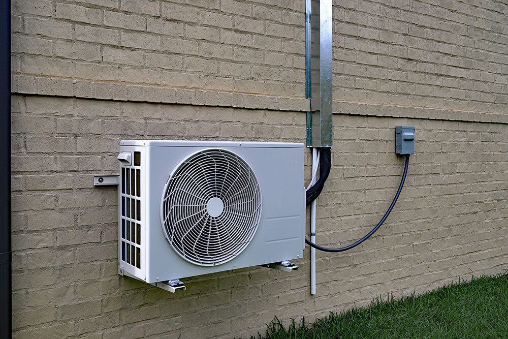 Outdoor air conditioning unit mounted on a brick wall, with connected cables leading upward and grass at the base.