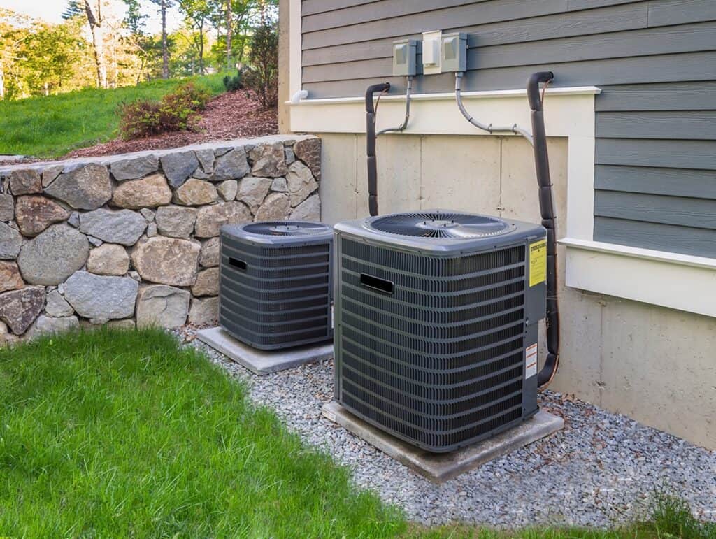 Two outdoor air conditioning units are installed on concrete slabs beside a house, with a stone wall and grassy area nearby.