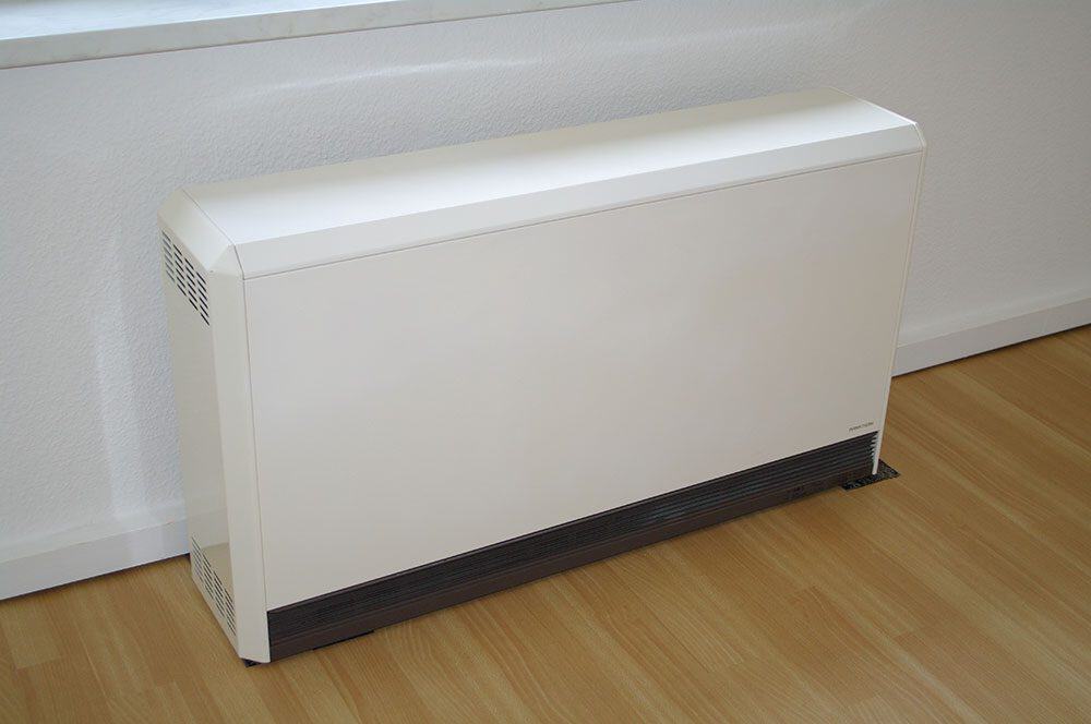 White electric convection heater placed against a wall on a wooden floor in a room.