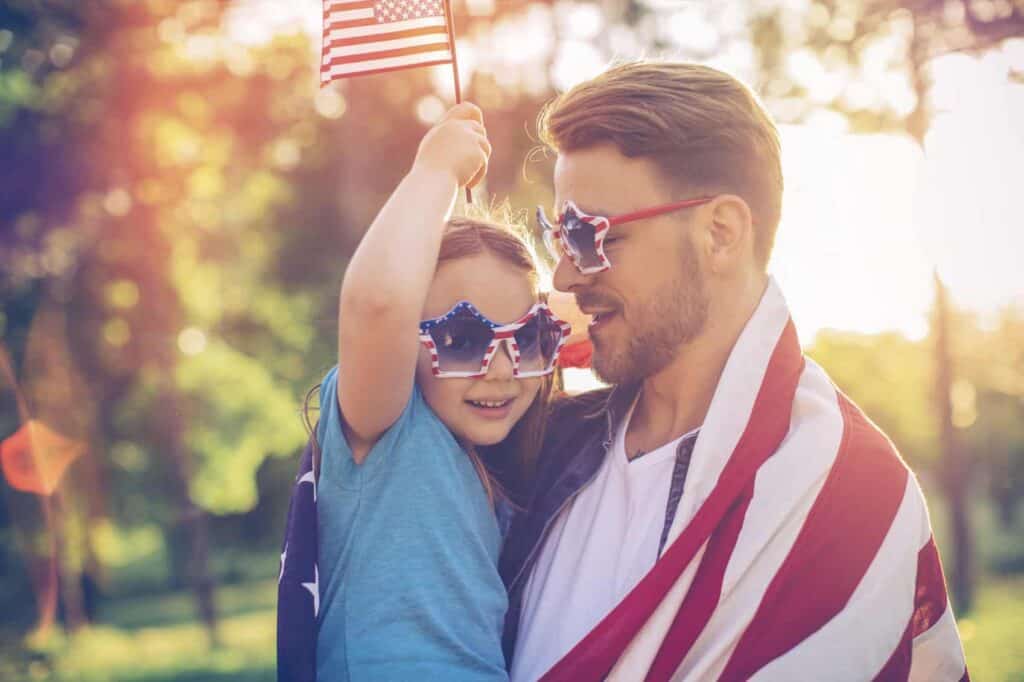 Man and child, both wearing star-shaped sunglasses, hold and are draped in American flags. They are outdoors with sunlight filtering through trees.