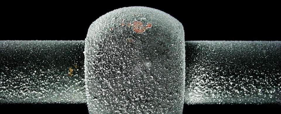 Close-up of a frost-covered metal pipe against a black background, showing a central round connector with visible rust patches.