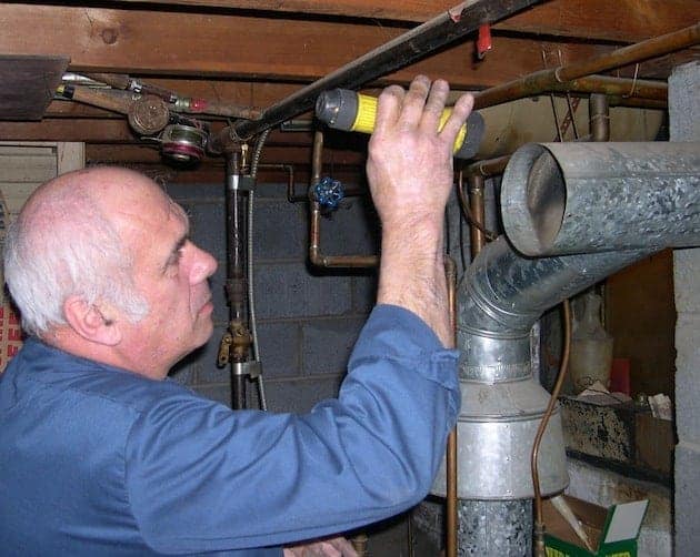 An older man in a blue shirt inspects plumbing in a basement, holding a flashlight to illuminate pipes and ducts.