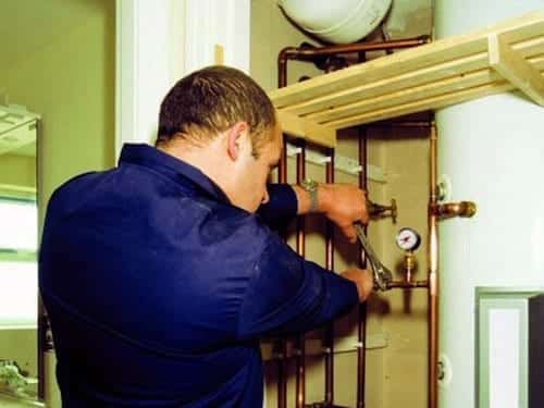 A person in blue overalls is repairing or adjusting pipes near a boiler tank, using wrench tools.