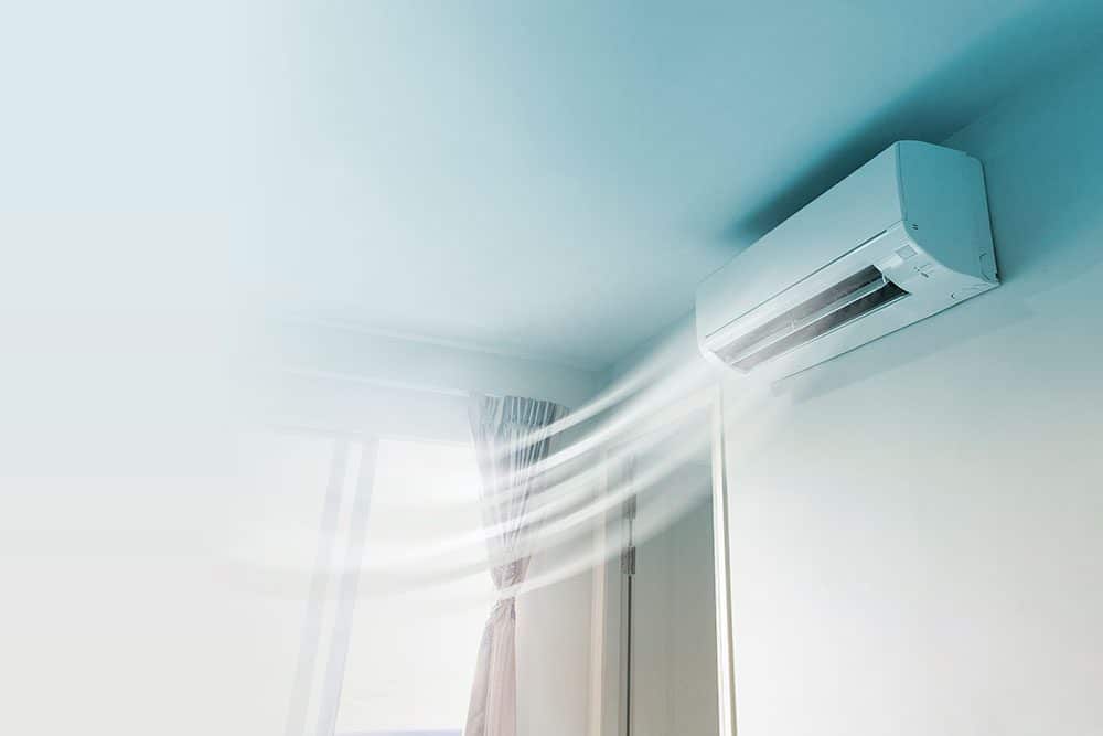 Wall-mounted air conditioner blowing cool air in a bright room with a window and curtain partially visible.