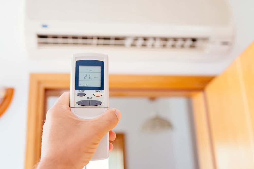 A hand holding a remote control set to 21°C, pointed at a wall-mounted air conditioner—showing how to use an AC for fall or cooler weather in the room.