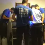 Four people in blue shirts examining equipment in a dimly lit room. One person's shirt reads "Never Homes Happy.
