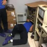 Person is lying on the floor with their head inside a cabinet, surrounded by shelves and boxes. Another person stands nearby.