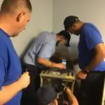 Four men in blue shirts and caps working on cables and equipment in a corner of a room.