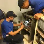 Three technicians in blue shirts and caps are examining and working on machinery in a small room, with one technician using a tool inside the equipment.