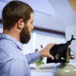 A bearded man in a blue shirt examines a small object on a stand in an indoor setting.