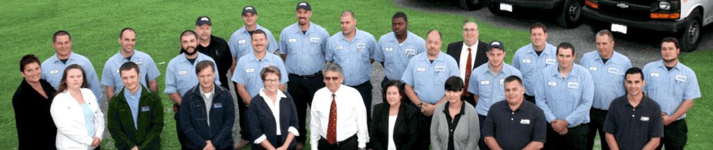 A group of people in uniforms and business attire pose outdoors on grass with vehicles in the background.