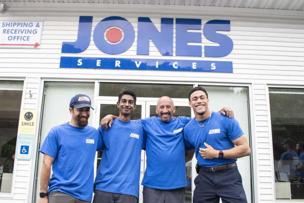 Four men in blue shirts with "Jones Services" text stand smiling in front of a building with a "Jones Services" sign.