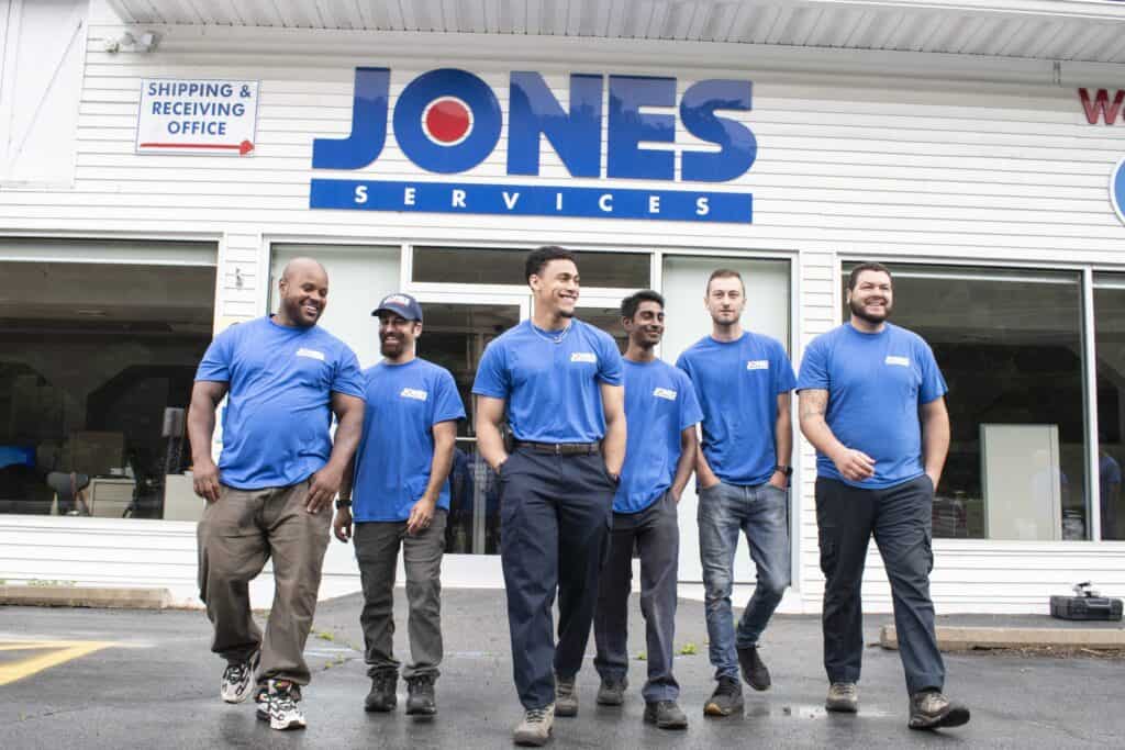 Six men in blue uniforms stand and walk in front of a building labeled "Jones Services," with a sign for "Shipping & Receiving Office" visible.