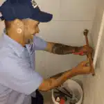 Person in a blue cap and shirt using a wrench on plumbing in a tiled area, with a bucket containing tools nearby.