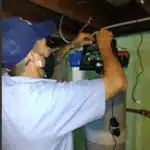 Person in blue uniform and cap working with electronic equipment in a room with exposed wiring.