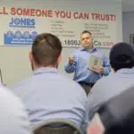 A man in a blue shirt speaks to an audience in a room with a banner reading "Tell Someone You Can Trust!" and contact information.