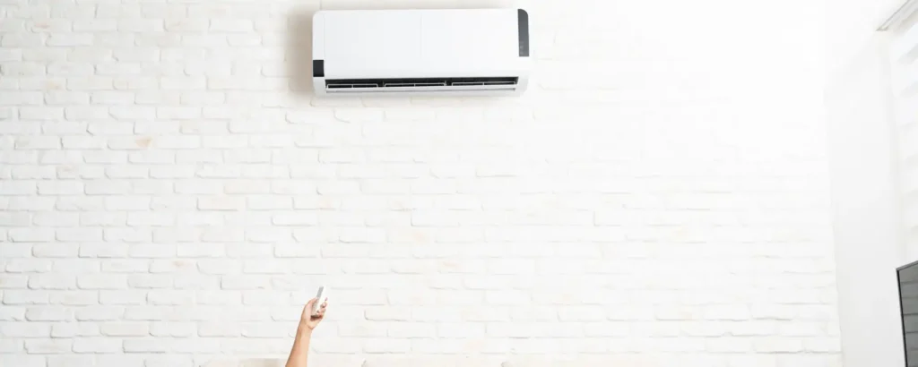 A hand holding a remote points towards a wall-mounted air conditioning unit on a white brick wall.