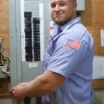 Person in a light blue uniform adjusting an electrical panel, with tools nearby, and an American flag patch on their sleeve.
