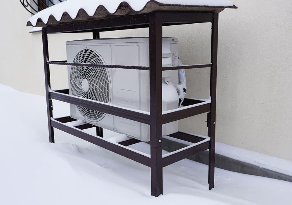 An outdoor air conditioning unit is partially covered in heavy snow, sheltered by a metal frame next to a building wall to help protect the heating system.