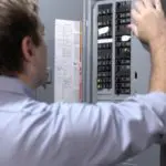 Person adjusting breakers in an electrical panel, wearing a light blue shirt, standing indoors.